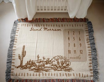 May include: A beige woven baby milestone blanket with a desert landscape design in brown. The blanket has a fringe border with red, blue, and gray threads. The name "David Morrison" is printed on the blanket, along with a section for tracking baby's age in months.