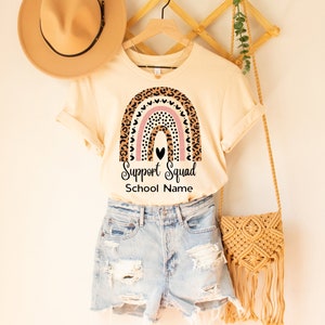 May include: A cream colored t-shirt with a rainbow design featuring leopard print and pink hearts. The text "Support Squad" and "School Name" is printed below the rainbow.