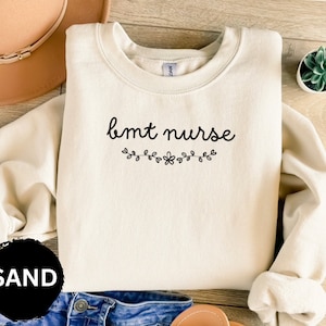 May include: A cream-colored sweatshirt with the text "bmt nurse" in black cursive lettering. The text is accented with a small floral design. The sweatshirt is labeled "SAND".