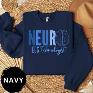 May include: Navy blue sweatshirt with the word "NEUR" in blue block letters and a brain graphic. Below the word "NEUR" is the text "EEG Technologist" in a script font. A black circle with the word "NAVY" is in the lower left corner.