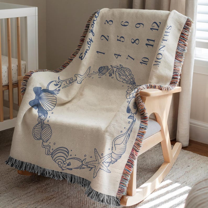 May include: Cream-colored baby milestone blanket with a blue seashell design and fringe trim. The blanket features the name "Emily Jones" and numbers 1-12 with the word "MONTHS". The blanket is draped over a wooden rocking chair.