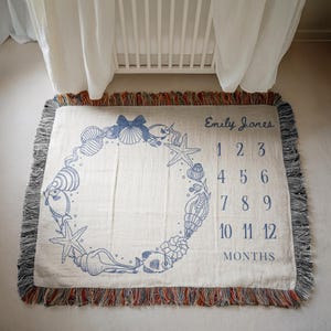 May include: Cream-colored woven baby milestone blanket with a blue seashell and starfish design. The blanket has a fringed edge and includes the name "Emily Jones" and numbers 1-12 for monthly photos.
