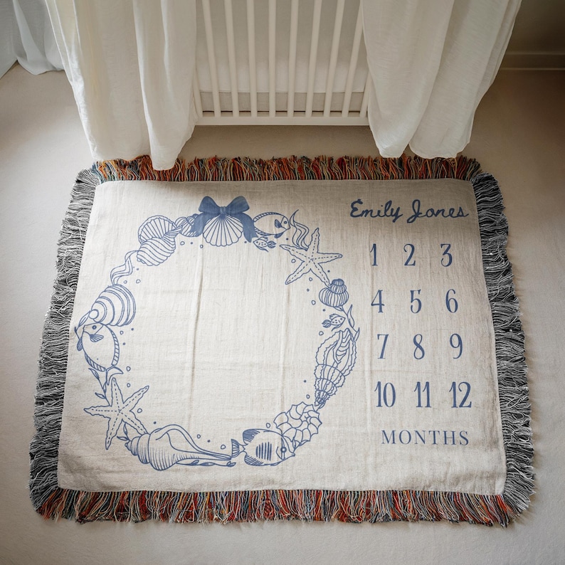 May include: Cream-colored woven baby milestone blanket with a blue seashell and starfish design. The blanket has a fringed edge and includes the name "Emily Jones" and numbers 1-12 for monthly photos.