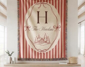 May include: A personalized throw blanket with a red and cream striped pattern. The center features an oval design with the letter "H", the name "The Hawkins", and a cabin scene. The blanket has a fringed edge.