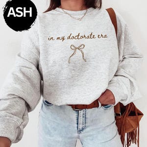 May include: A light gray sweatshirt with the text "in my doctorate era" and a bow graphic in brown. The sweatshirt has long sleeves and a crew neck. The word "ASH" is in a black circle in the upper left corner.