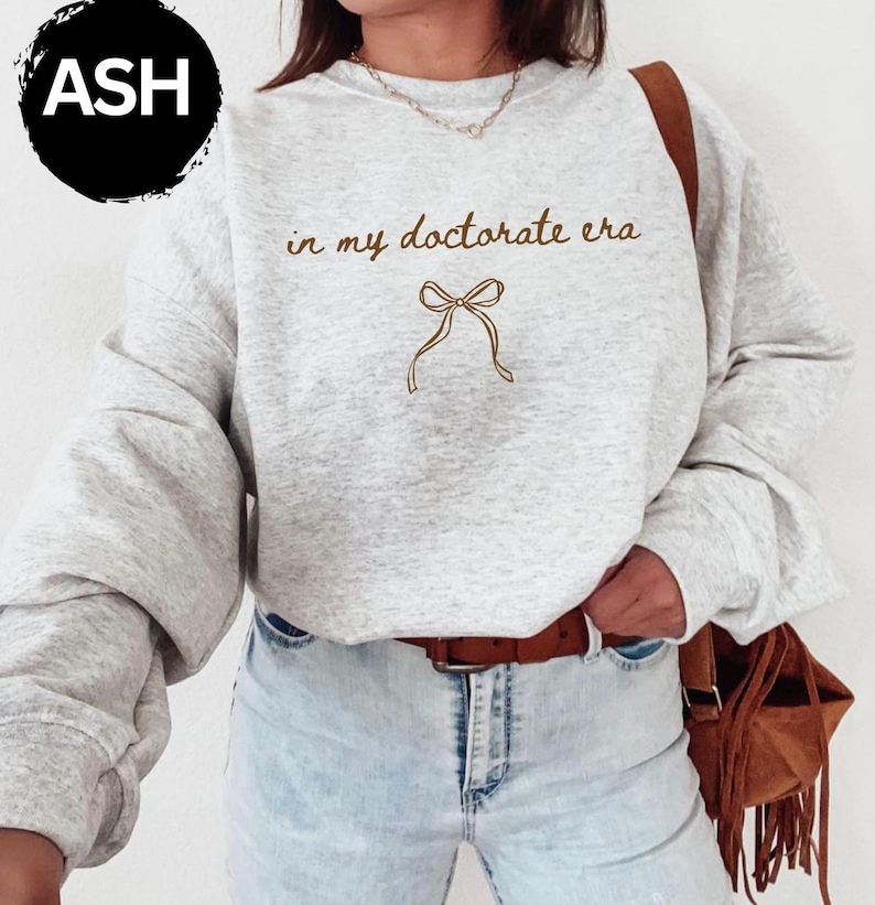 May include: A light gray sweatshirt with the text "in my doctorate era" and a bow graphic in brown. The sweatshirt has long sleeves and a crew neck. The word "ASH" is in a black circle in the upper left corner.