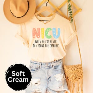 May include: A cream colored t-shirt with the text "NICU When you're never too young for caffeine" in rainbow colors. The shirt is hanging on a wooden hanger with a brown hat and a woven bag.