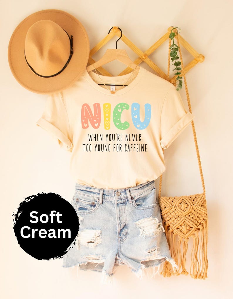 May include: A cream colored t-shirt with the text "NICU When you're never too young for caffeine" in rainbow colors. The shirt is hanging on a wooden hanger with a brown hat and a woven bag.