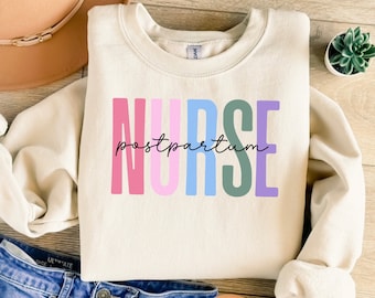 May include: A light beige sweatshirt with the word "NURSE" printed in colorful letters. The word "postpartum" is written in black script below the word "NURSE".