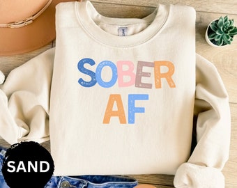 May include: A cream-colored sweatshirt with the text "SOBER AF" in colorful letters. The letters are in a distressed font and have a vintage look.