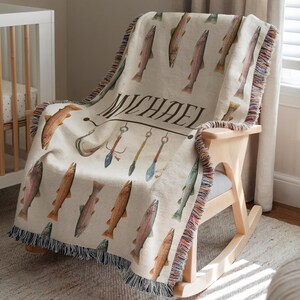 May include: A cream-colored throw blanket with a repeating pattern of watercolor fish. The blanket features the name "MICHAEL" and fishing-themed illustrations. The edges are finished with a fringed trim, and the blanket is draped over a wooden rocking chair.