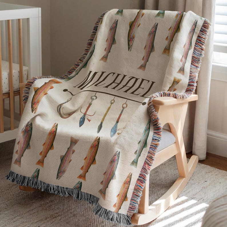 May include: A cream-colored throw blanket with a repeating pattern of watercolor fish. The blanket features the name "MICHAEL" and fishing-themed illustrations. The edges are finished with a fringed trim, and the blanket is draped over a wooden rocking chair.