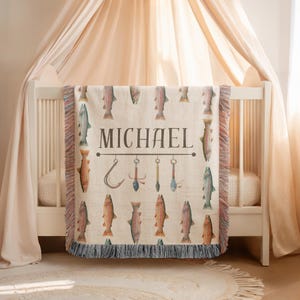 May include: A beige throw blanket with a fishing theme, featuring watercolor illustrations of fish and fishing tools. The name "MICHAEL" is printed in large, dark letters. The blanket has a fringed edge and is draped over a white crib.