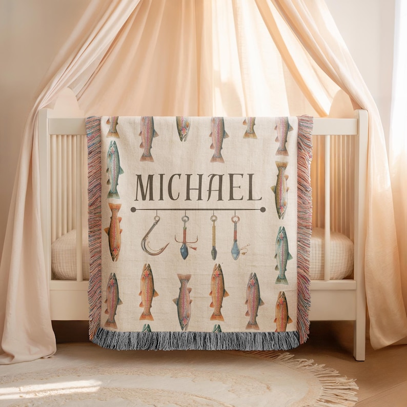 May include: A beige throw blanket with a fishing theme, featuring watercolor illustrations of fish and fishing tools. The name "MICHAEL" is printed in large, dark letters. The blanket has a fringed edge and is draped over a white crib.
