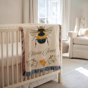 May include: A beige baby blanket with a large watercolor bumblebee design. The blanket has a fringed edge and features the name "Olivia Jones" with floral and honeycomb accents. The blanket is draped over a white crib.