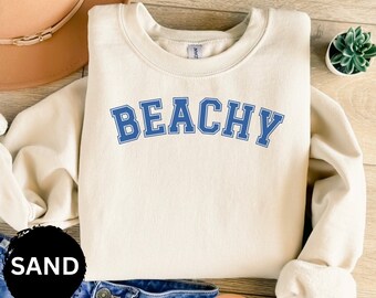 May include: A cream colored sweatshirt with the word "BEACHY" in blue block letters. The sweatshirt is laying on a wooden surface with a brown hat and a potted plant.