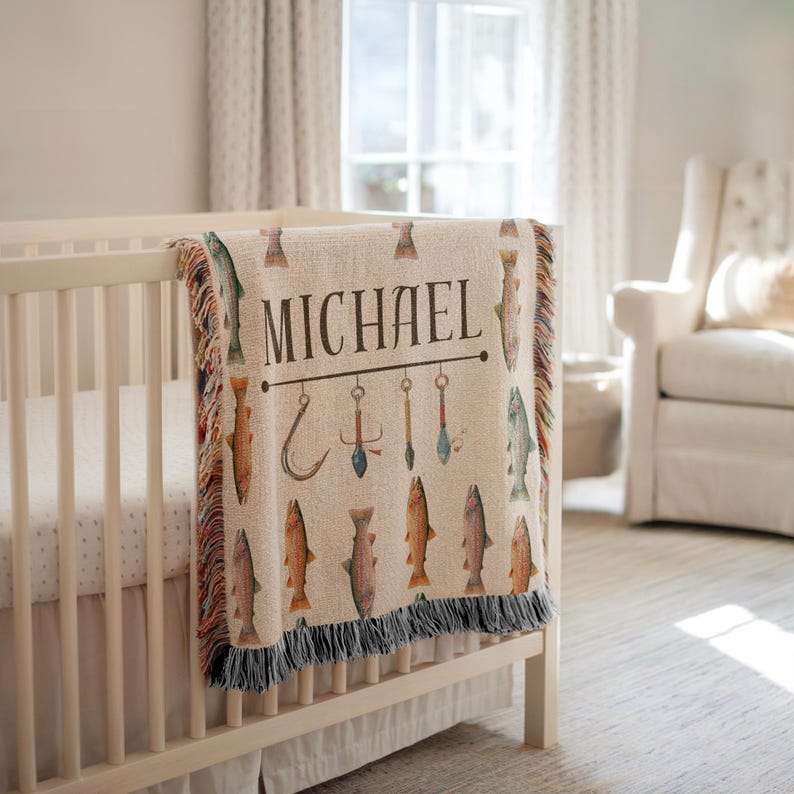 May include: A beige baby blanket with a fishing theme, draped over a crib. The blanket features the name "MICHAEL" in large letters, along with illustrations of fish, hooks, and fishing tools. The blanket has a fringed edge.