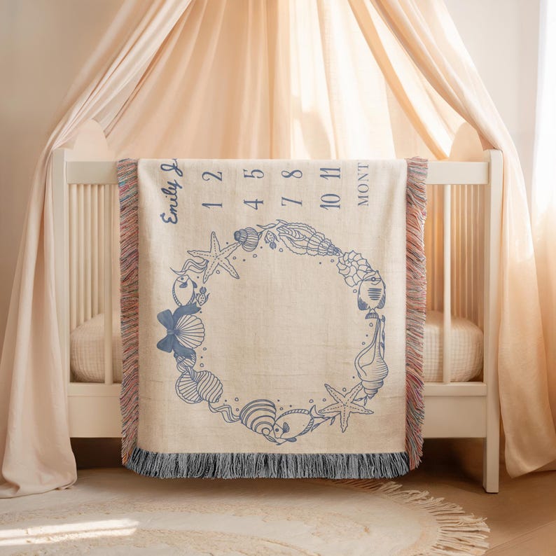 May include: A beige baby milestone blanket with a blue seashell design. The blanket has a fringe border and features the name "Emily J" and numbers indicating months. The design includes starfish, shells, and other sea creatures.