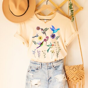 May include: A cream colored t-shirt with a graphic of colorful hummingbirds and flowers. The shirt is hanging on a wooden hanger and is paired with denim shorts.