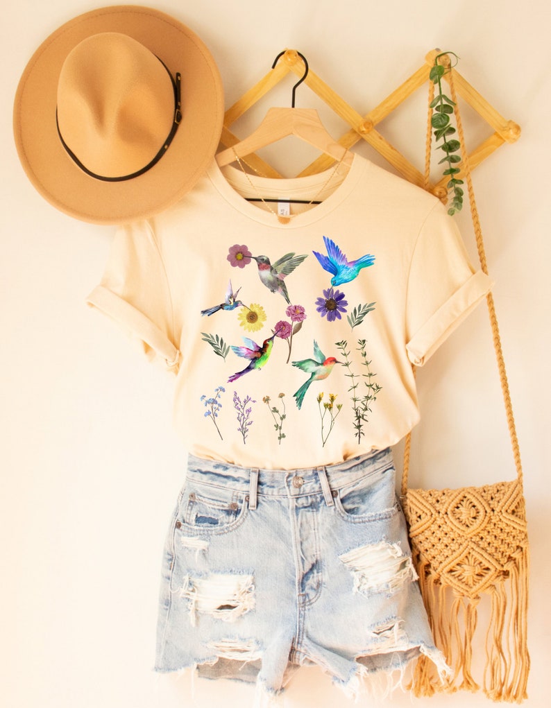 May include: A cream colored t-shirt with a graphic of colorful hummingbirds and flowers. The shirt is hanging on a wooden hanger and is paired with denim shorts.