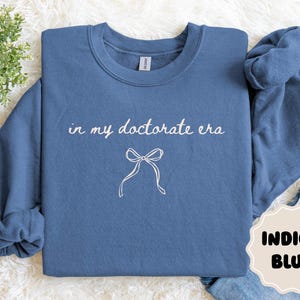 May include: Indigo blue sweatshirt with the text "in my doctorate era" and a bow graphic. The sweatshirt is a crew neck style. The words and bow are in white. The color of the sweatshirt is indigo blue.