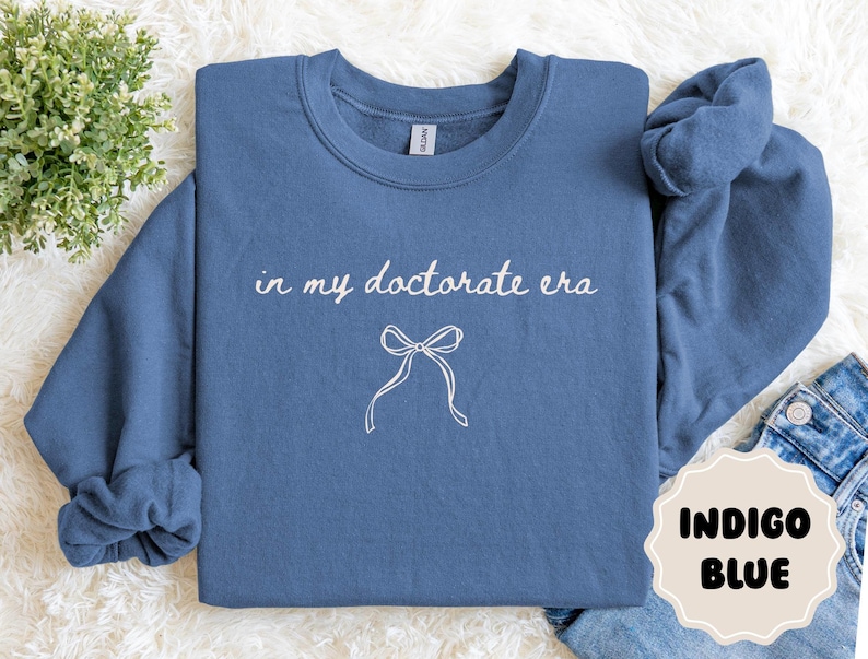 May include: Indigo blue sweatshirt with the text "in my doctorate era" and a bow graphic. The sweatshirt is a crew neck style. The words and bow are in white. The color of the sweatshirt is indigo blue.