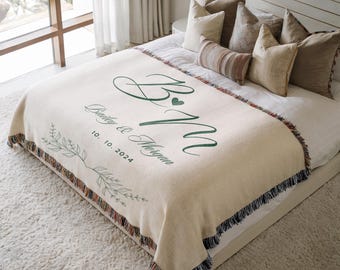May include: Cream-colored woven blanket with green lettering and a floral design. The blanket features the initials "B & M" with a heart, the names "Bailey & Morgan", and the date "10.10.2024". The blanket has a fringed edge.