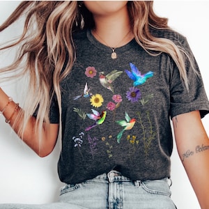 May include: A dark gray t-shirt with a colorful hummingbird and floral print. The shirt has a crew neck and short sleeves.