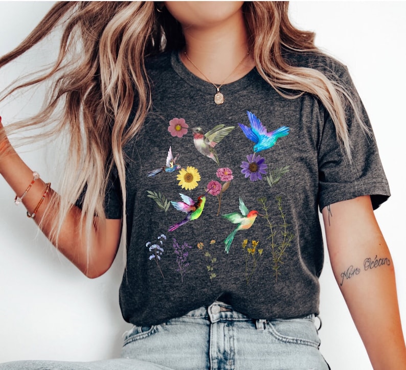 May include: A dark gray t-shirt with a colorful hummingbird and floral print. The shirt has a crew neck and short sleeves.