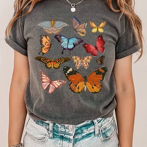 K&ouml;nnte beinhalten: Ein dunkelgraues T-Shirt mit einem bunten Schmetterlingsdruck. Die Schmetterlinge sind in verschiedenen Blaut&ouml;nen, Rot, Orange, Gelb und Rosa.