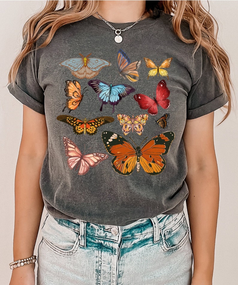 K&ouml;nnte beinhalten: Ein dunkelgraues T-Shirt mit einem bunten Schmetterlingsdruck. Die Schmetterlinge sind in verschiedenen Blaut&ouml;nen, Rot, Orange, Gelb und Rosa.