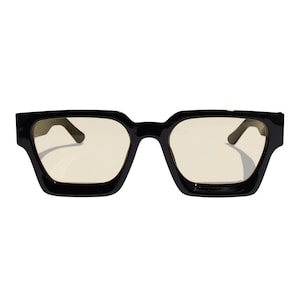 Op de afbeelding: Zwarte vierkante bril met lichtgele glazen. De bril heeft een dik, gedurfd montuurontwerp. De glazen zijn lichtgeel van kleur, wat een contrast vormt met het zwarte montuur. Een modern brillenaccessoire.