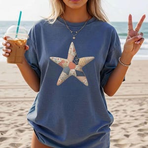 Pode incluir: Camiseta azul com um grande design de estrela feito de tecidos de patchwork. A estrela incorpora padrões florais, listrados e geométricos. A camiseta é de corte folgado, adequada para uso casual. A imagem é tirada numa praia.
