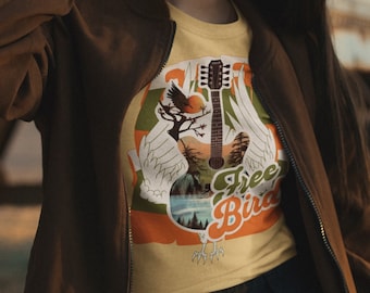 Könnte beinhalten: Ein gelbes T-Shirt mit einem Grafikdruck einer akustischen Gitarre in Vogelform mit Flügeln, einem Sonnenuntergang, Bäumen und dem Text Free Bird. Das T-Shirt wird unter einer braunen Reißverschlussjacke mit einer schwarzen Hose getragen.