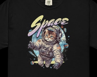 May include: A black short-sleeve t-shirt featuring a graphic of a tabby cat wearing a white astronaut suit. Above the cat, the word Space is printed in a yellow and purple gradient font. The background includes blue planets and stars.
