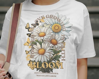 May include: A white short-sleeved t-shirt featuring a graphic of daisies, butterflies, and the text "LOVE GROWS IN FAITH", "BLOOM", and "CULTIVATE KINDNESS & WATCH LOVE GROW". The shirt is worn with blue jeans and a brown shoulder bag.
