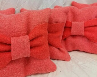 May include: A collection of coral-coloured pillows with a felted texture, each adorned with a contrasting red bow. The pillows are made of a soft, felted material. The arrangement is in a row.