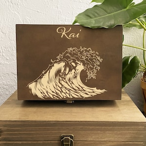 Könnte beinhalten: Eine braune Holzkiste mit einem gelaserten Wellenmuster und dem Namen "Kai" in eleganter Schrift. Die Kiste steht auf einer größeren, ähnlichen Holzkiste. Das Design ist hellbraun.