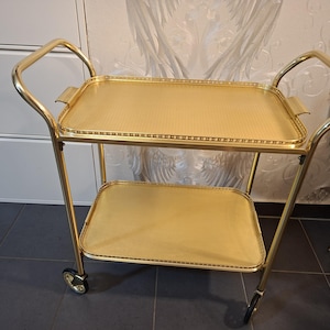 May include: A two-tiered gold-toned metal serving cart featuring a rectangular frame with rounded corners, two handles, and four wheels. Each tier consists of a flat tray with a textured surface and a decorative perforated edge border.