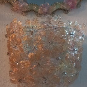 May include: A square wall sconce featuring a floral design made of clear and peach-coloured glass flowers. The sconce has small, sparkling accents and is mounted on a white wall. A decorative mirror is above the sconce.
