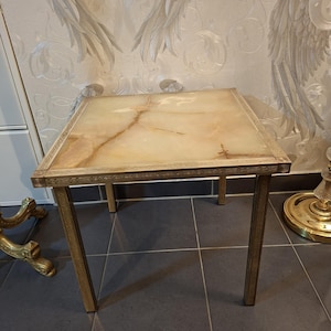 May include: A small, square side table with a light-colored, marble-like top and gold-colored metal frame. The table has four straight legs and decorative detailing around the top edge. The table is approximately 24 inches tall.