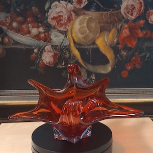 Könnte beinhalten: Eine leuchtend orangefarbene Glasschale mit einem einzigartigen, abstrakten Design. Die Schale wird auf einem schwarzen Drehteller präsentiert.