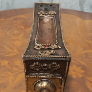 Puede incluir: Un quemador de incienso de oro y cobre con intrincados diseños florales y una pequeña joya roja. El quemador tiene una forma rectangular y se encuentra sobre una superficie de madera.