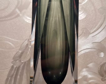 May include: A clear glass vase with a geometric shape and a dark green glass insert. The vase has a faceted design and a pointed base.
