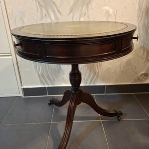 May include: A dark brown wooden drum table with an oval top featuring a green leather inlay with gold-coloured border detailing. The table has a central pedestal base with three curved legs ending in metal claw feet and two small side drawers.