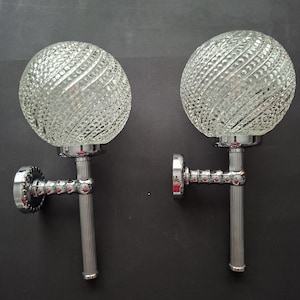 May include: A pair of wall sconces featuring spherical, clear textured glass globes with a diagonal ribbed pattern. Each light fixture has a polished chrome metal base, a vertical fluted arm, and a circular wall mounting plate with a decorative edge.