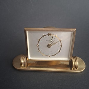Puede incluir: Un reloj de escritorio rectangular de tono dorado con esfera plateada, que presenta el texto EUROPA y 4 JEWELS. El reloj tiene un marco dorado y se asienta sobre una base ovalada alargada con detalles acanalados, manecillas e índices dorados.