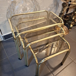 Puede incluir: Un juego de tres mesas nido con estructura de metal dorado y tapas de cristal transparente. Las mesas son rectangulares con esquinas redondeadas. Las patas son curvas y las mesas están dispuestas en forma de nido.