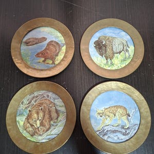 May include: A set of four circular coasters featuring a metallic brass-toned rim and a central illustration. The designs depict a beaver in water, a bison in a grassy field, a brown bear with a cub, and a lynx standing on a rocky surface.