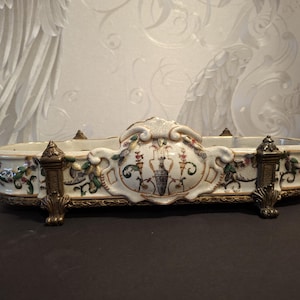 Op de afbeelding: Een langwerpige keramische schaal als tafelstuk met een crèmekleurige basis, versierd met complexe bloemmotieven en een centrale urnafbeelding. De schaal rust op sierlijke bronzen metalen voetjes en heeft bijpassende decoratieve handgrepen.
